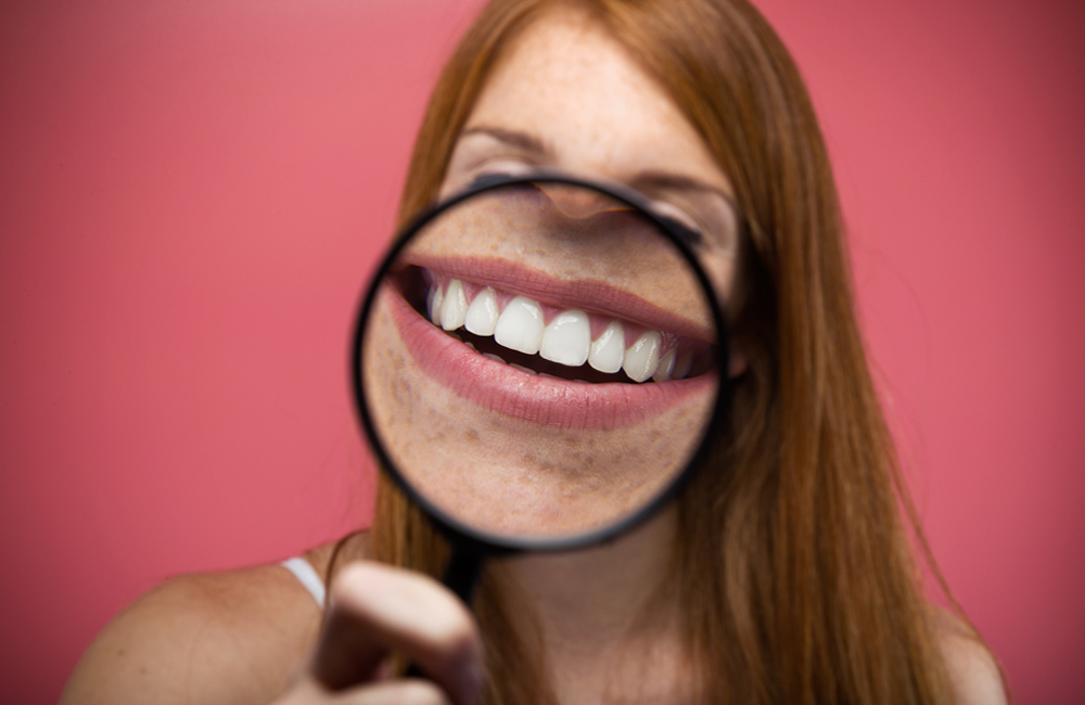 Pretty young woman showing teeth trought magnifying glass over pink background.
